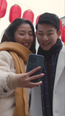 Video-Vertical-De-Una-Joven-Pareja-Asiática-De-Vacaciones-Posando-Para-Un-Selfie-En-Un-Teléfono-Móvil-En-Chinatown,-Londres,-Reino-Unido